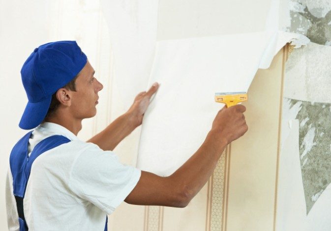 Pintor retirando papel pintado antiguo de una pared con espátula antes de colocar el nuevo